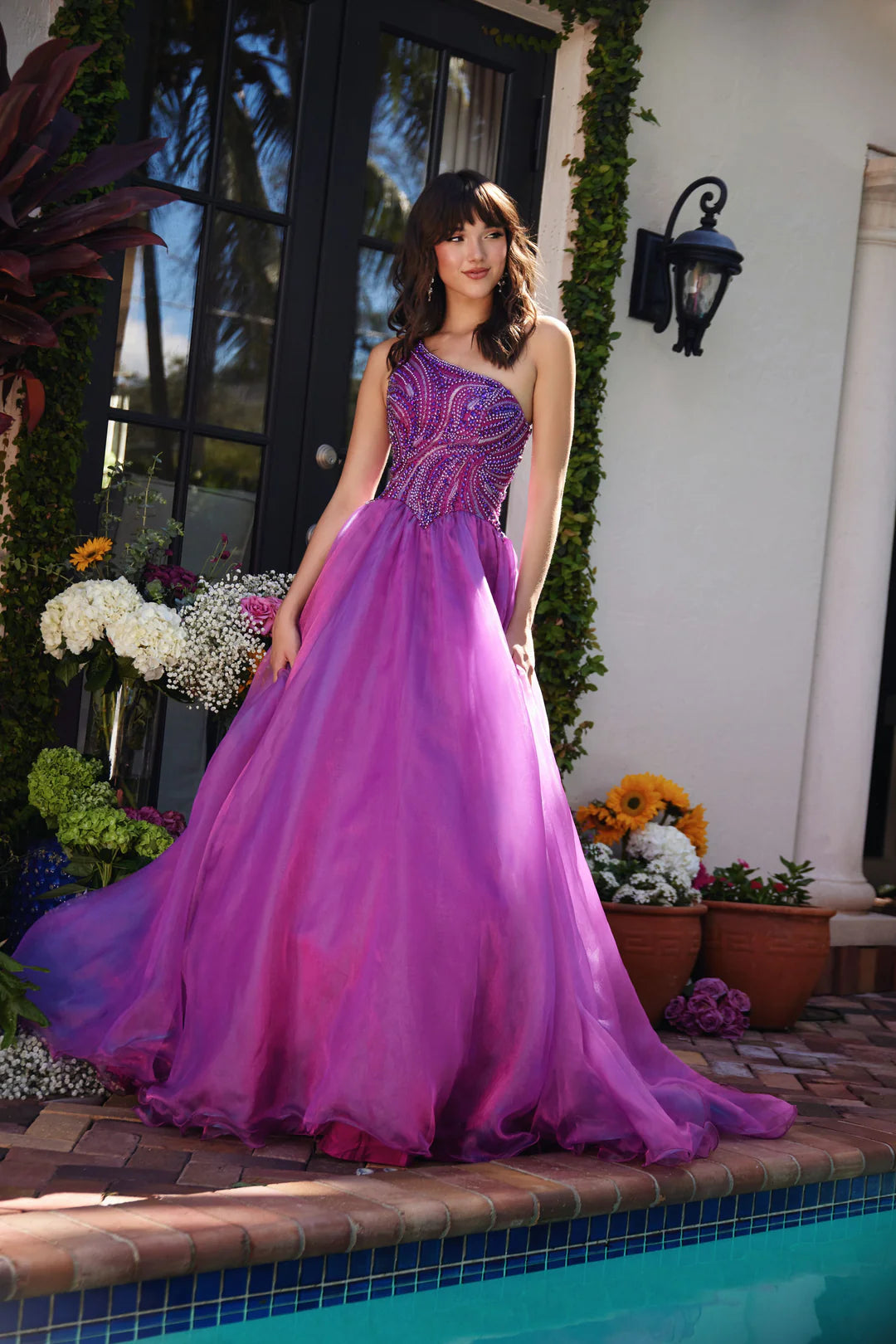 This Ava Presley gown, style 29258, exudes a captivating elegance with its magenta hue and one-shoulder design. The asymmetrical neckline adds a modern touch, while the fitted bodice features intricate beadwork in swirling patterns that accentuate the curves and create a sparkling effect.&nbsp;The full skirt is crafted from layers of lightweight fabric that flow gracefully, adding movement and volume to the gown. This design is perfect for formal events like proms, pageants, or gala evenings, offering a ble