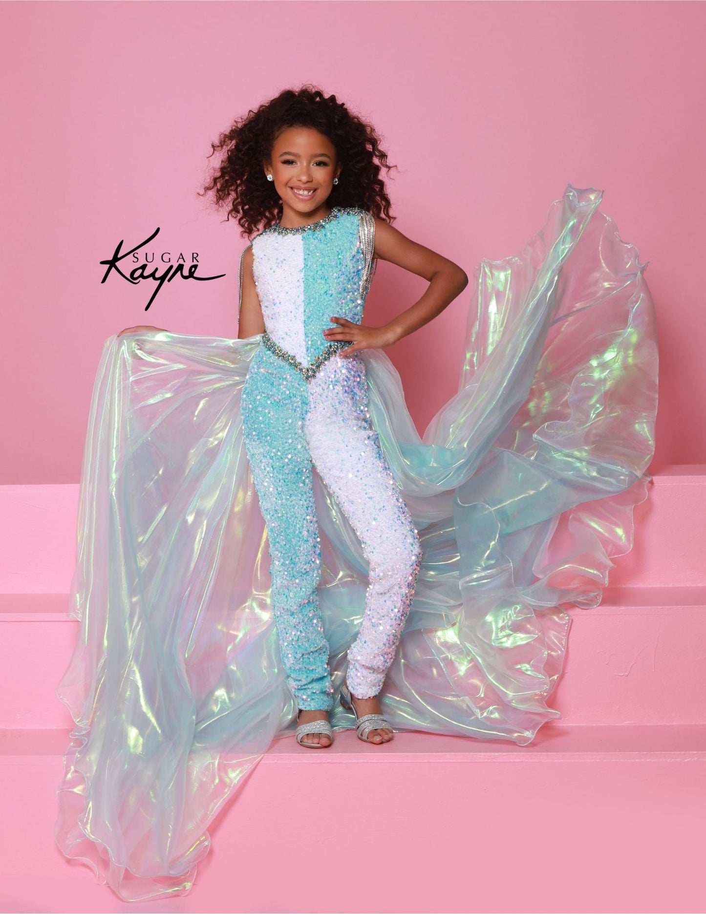 Get your little one ready to stand out in style with Sugar Kayne's C386 Girls Velvet Sequin Pageant Jumpsuit. This fun and fashionable piece features a stunning velvet fabric with shimmering sequin details, perfect for any pageant or special occasion. The fringe overskirt adds a touch of playful flair, making your child the center of attention. This sequin stretch velvet two-tone jumpsuit features crystal embellishments and beaded shoulder fringe. An iridescent organza cape flows from the waist, adding a dr