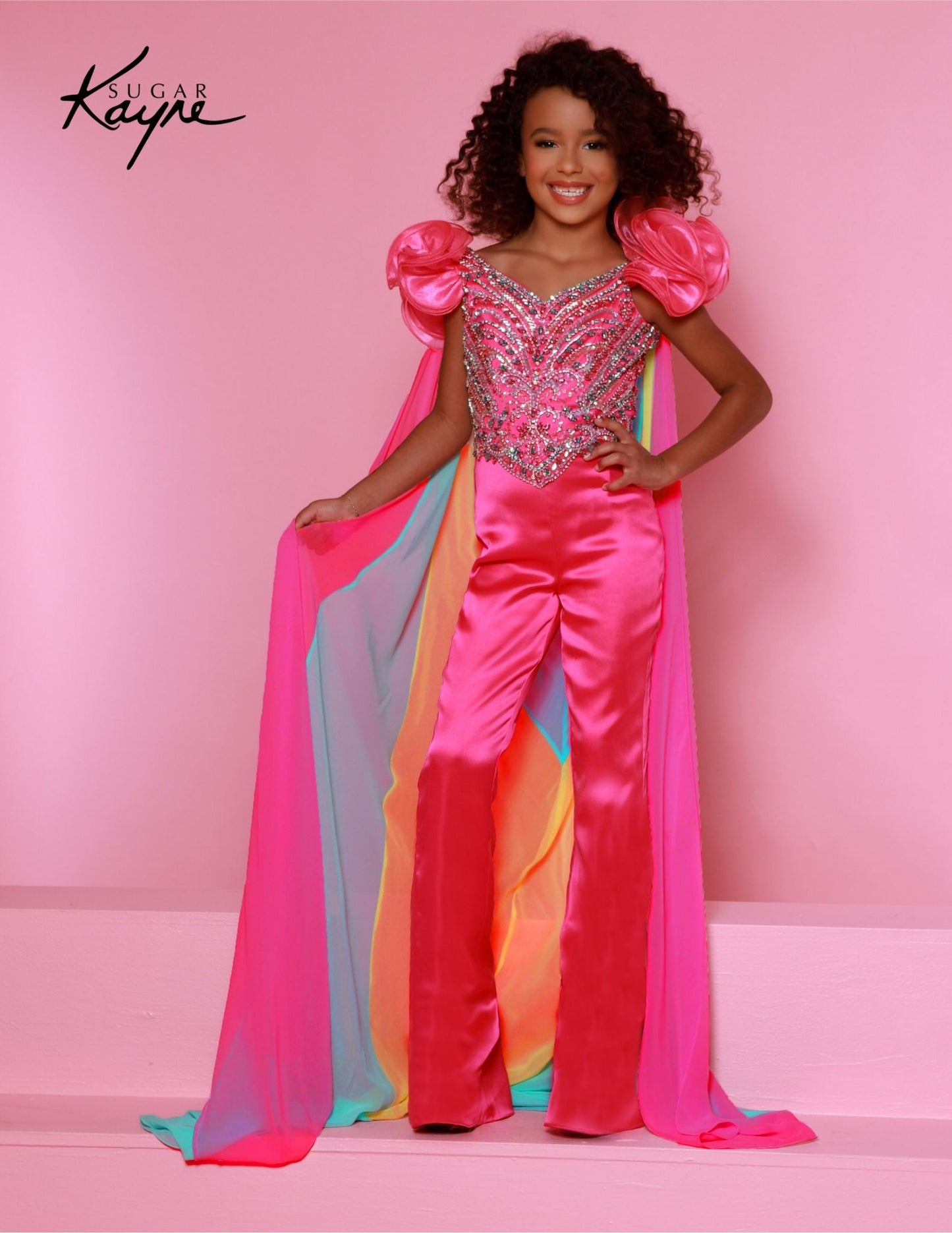 The Sugar Kayne C335 Girls Two Piece Fun Fashion Jumpsuit features a beaded bodice, a detachable cape, and a stylish, comfortable fit. Perfect for pageants, the outfit is sure to make your child look her best. Step into a whirlwind of whimsy with this Crepe Back Satin Jumpsuit. The beaded bodice, ruffle sleeves, and detachable colorful chiffon cape make every day a playful and stylish adventure.